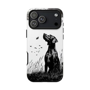 May include: A phone case featuring a black and white illustration of a dog looking up at birds in flight. The dog is sitting in tall grass, with a white background. The case has a black camera module.