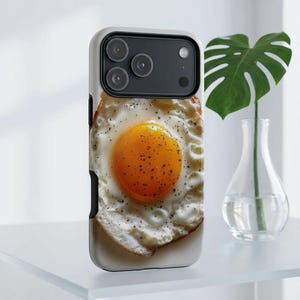 May include: A smartphone case featuring a high-resolution print of a fried egg with a bright orange yolk and white edges, seasoned with black pepper. The case has a black camera cutout area and a white border, designed for a triple-lens phone.
