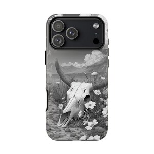 May include: A black and white phone case featuring a detailed illustration of a cow skull with large horns, surrounded by wildflowers and tall grass. The background depicts a landscape with mountains and clouds.