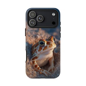 May include: A phone case featuring a detailed image of a frog with orange and brown markings, emerging from sand. The case has a dark blue border and a camera cutout.
