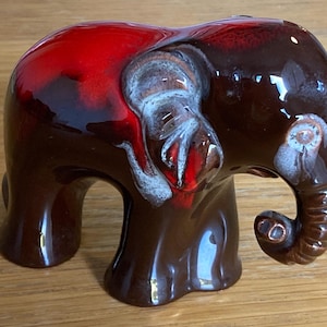 May include: A ceramic elephant ornament with a glossy finish. The elephant is mainly brown with a red top and white accents on the ears. The trunk is curled, and the ornament has a vintage aesthetic.