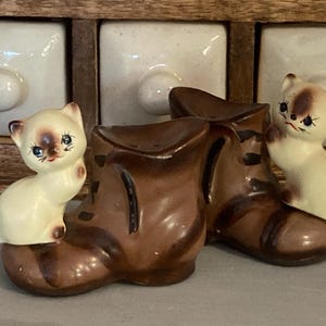 May include: A pair of ceramic salt and pepper shakers shaped like brown boots, each featuring a small cream-coloured kitten with dark brown ears, a dark brown face mask, and blue eyes perched on the side of the boot. The kittens have painted whiskers.