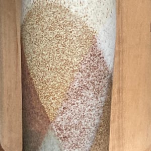 May include: A tall, cylindrical ceramic vase featuring a speckled, off-white upper section and a lower portion decorated with geometric, triangular colour blocks in shades of muted tan, earthy brown, and a soft, pale blue-grey with a matte finish.
