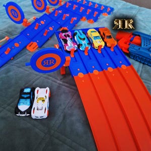 May include: A blue and orange toy race track set with four toy cars, two of which are blue and two of which are orange. The track has a ramp and a target. The set also includes a blue and black toy gun.