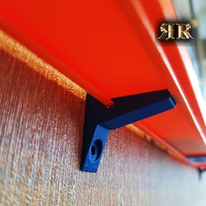 May include: A close-up of a red shelf bracket mounted to a wooden wall. The bracket is made of metal and has a black finish. The shelf is not visible in the image.
