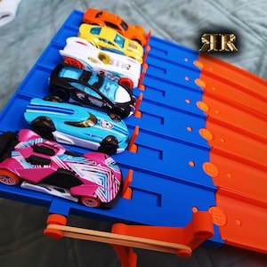Könnte beinhalten: Eine blaue und orangefarbene Hot Wheels-Rennstrecke mit sechs bunten Spielzeugautos an der Startlinie. Die Autos sind orange, gelb, weiß, schwarz, hellblau und rosa. Die Rennstrecke befindet sich auf einer hellblauen Oberfläche.