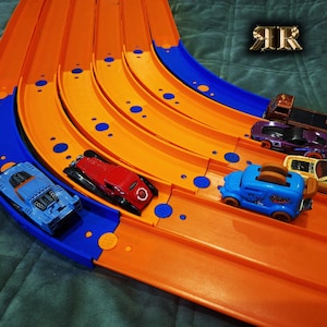 以下が含まれることがあります： オレンジと青のレーンがあるおもちゃのレーストラック。 青、赤、紫、黄色のモデルなど、いくつかのカラフルなおもちゃの車がトラック上に配置され、レースの準備ができています。 トラックはカーブと傾斜があり、ダイナミックな遊び環境を示唆しています。