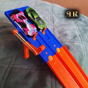 Könnte beinhalten: Ein blaues und oranges Hot Wheels Rennbahnset mit zwei Spielzeugautos, eines pink und eines grün, auf der Strecke.