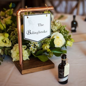 May include: A white table card with black text that reads "The Bolingbroke" is hanging from a copper pipe stand. The stand is on a white tablecloth with a bouquet of white and green flowers in front of it. There are several small bottles of liquor on the table.