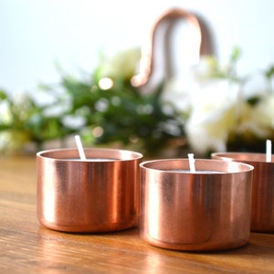 May include: Three copper candle holders with white candles on a wooden surface. The candle holders are round and have a shiny finish.