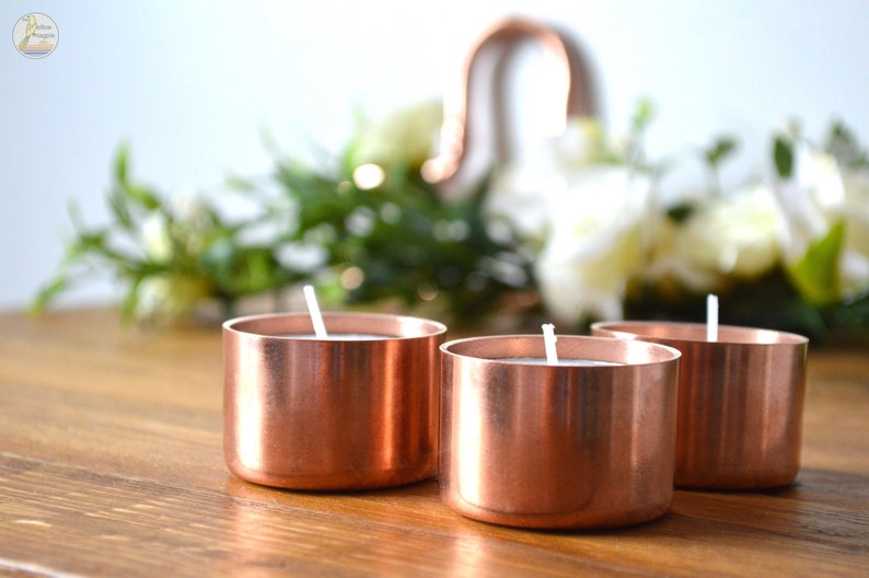 May include: Three copper candle holders with white candles on a wooden surface. The candle holders are round and have a shiny finish.