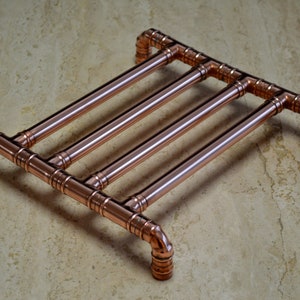 May include: Copper pipe trivet with a rectangular design. The trivet is made of copper pipes and fittings, with four parallel pipes supported by a frame. The copper has a shiny, metallic finish. The trivet is designed to protect surfaces from hot items.
