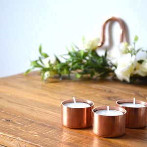 May include: Three copper candle holders with white tea lights on a wooden table.