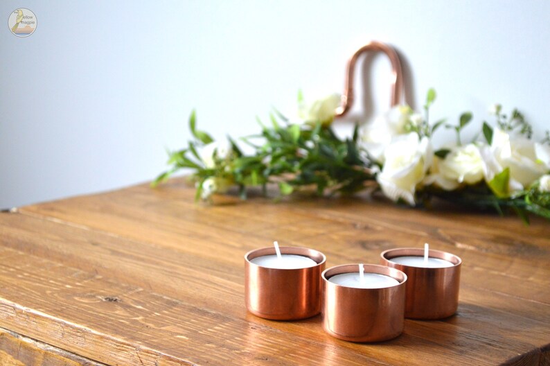 May include: Three copper candle holders with white tea lights on a wooden table.