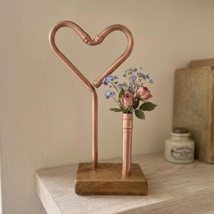 May include: A copper pipe sculpture shaped like a heart attached to a rectangular wooden base. Next to the heart, a vertical copper pipe segment acts as a vase, holding a small arrangement of dried pink roses and tiny blue forget-me-not flowers.