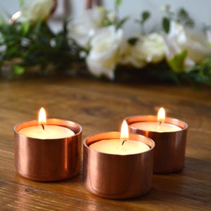 May include: Three copper candle holders with white tea lights burning inside. The candle holders are on a wooden surface.