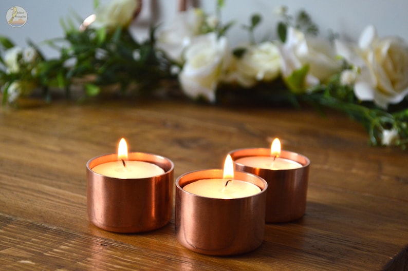 May include: Three copper candle holders with white tea lights burning inside. The candle holders are on a wooden surface.