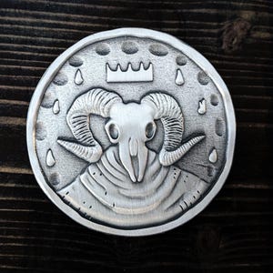 May include: A silver-colored round pendant featuring a ram skull with large curved horns, a crown, and teardrop shapes around the edge. The pendant has a detailed, textured surface and is likely made of metal.