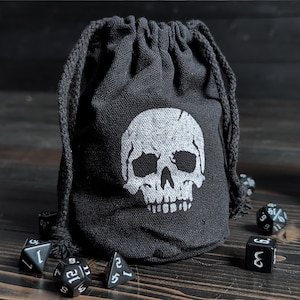 May include: Black drawstring dice bag with a white skull graphic. The bag is made of a textured fabric and is surrounded by several black dice with white numbering. The bag is closed with a black drawstring.