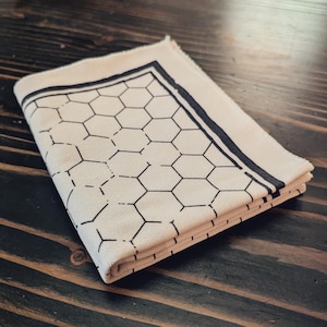 Pode incluir: Guardanapo de tecido dobrado creme com um padrão de favo de mel preto e uma borda preta. O guardanapo é feito de um tecido macio e é cuidadosamente dobrado, mostrando claramente o padrão. O design é moderno e geométrico.