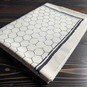 Puede incluir: Una pila de servilletas de tela plegadas, de color blanquecino. Cada servilleta presenta un patrón de panal de abeja negro y un borde negro. Las servilletas están apiladas sobre una superficie de madera oscura.