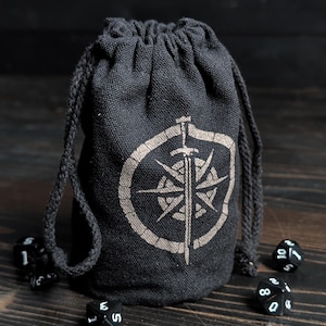 May include: Black drawstring dice bag with a compass rose and sword design in a light tan color. The bag is made of a textured fabric and is surrounded by several black dice with white numbers. The bag is closed with a black drawstring.
