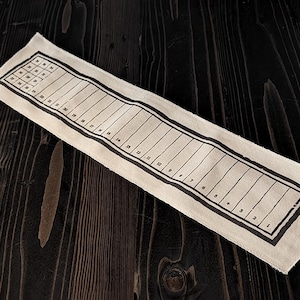 May include: A beige linen ruler with black printed markings and numbers. The ruler has a grid at one end and numbered sections along its length. The ruler is lying on a dark wooden surface.
