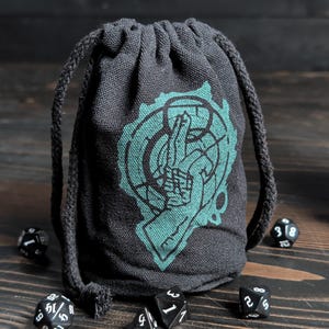 May include: A dark gray drawstring dice bag with a teal graphic of a hand and gear design. The bag is surrounded by several black dice with white numbering. The bag is made of a textured fabric.