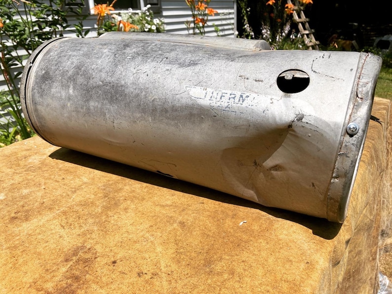 Op de afbeelding: Een zilveren metalen cilinder met een deuk en een klein gat aan de zijkant. De cilinder heeft de tekst "LURM" erop.