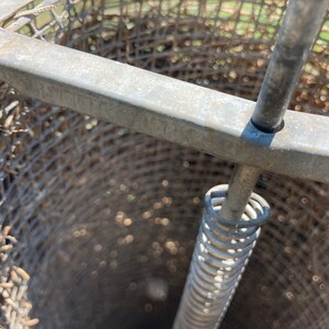 Op de afbeelding: Close-up van een metalen kooi met een veermechanisme. De kooi is gemaakt van gaas en heeft een ronde opening aan de bovenkant. De veer is bevestigd aan een metalen staaf die door het midden van de kooi loopt.