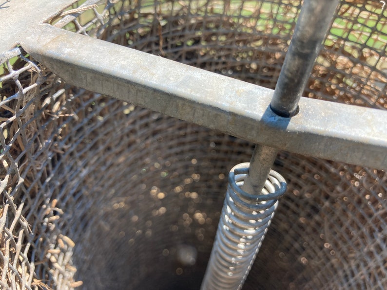 Op de afbeelding: Close-up van een metalen kooi met een veermechanisme. De kooi is gemaakt van gaas en heeft een ronde opening aan de bovenkant. De veer is bevestigd aan een metalen staaf die door het midden van de kooi loopt.