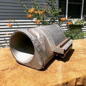Op de afbeelding: Een vintage metalen voorwerp met een grote, ronde opening en een kleinere, rechthoekige opening aan de zijkant. Het voorwerp is verweerd en heeft een roestige patina. Het ligt op een bruin, getextureerd oppervlak.
