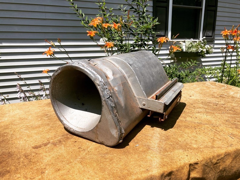 Op de afbeelding: Een vintage metalen voorwerp met een grote, ronde opening en een kleinere, rechthoekige opening aan de zijkant. Het voorwerp is verweerd en heeft een roestige patina. Het ligt op een bruin, getextureerd oppervlak.
