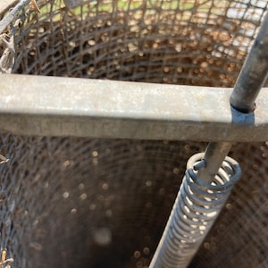 Op de afbeelding: Een close-up van een metalen honing extractor met een veerbelaste mechanisme. De extractor is gemaakt van een draadgaas en heeft een metalen handvat.