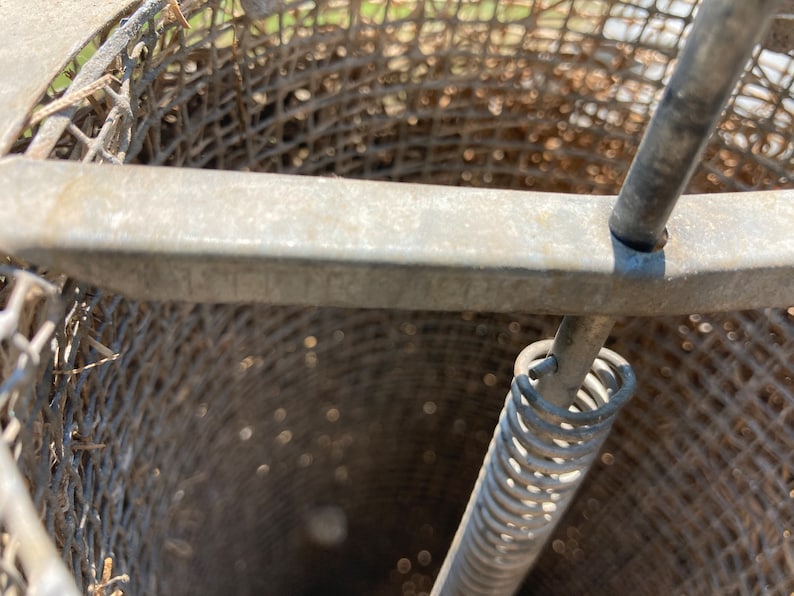 Op de afbeelding: Een close-up van een metalen honing extractor met een veerbelaste mechanisme. De extractor is gemaakt van een draadgaas en heeft een metalen handvat.