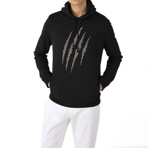 May include: Black hooded sweatshirt with a claw mark design. The claw marks are made of a shimmering, multi-colored material. The sweatshirt has a front pocket and a drawstring hood. The model is wearing white pants.