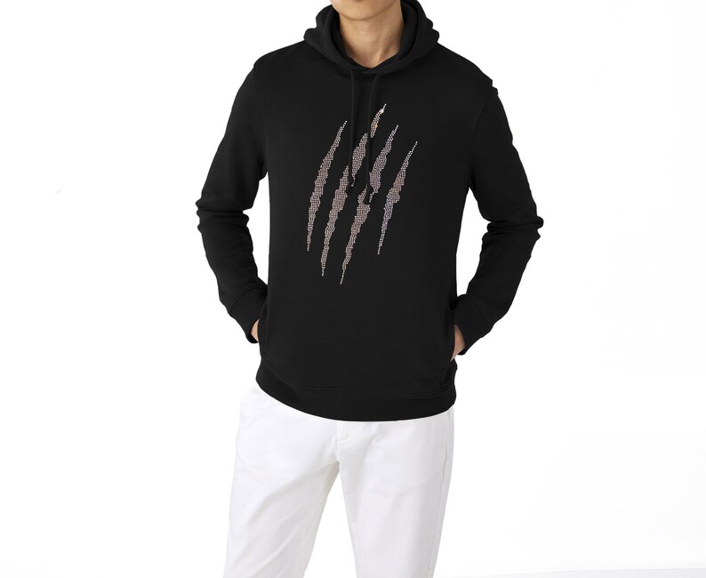 May include: Black hooded sweatshirt with a claw mark design. The claw marks are made of a shimmering, multi-colored material. The sweatshirt has a front pocket and a drawstring hood. The model is wearing white pants.