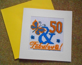May include: A white card with a blue and orange cross-stitch design. The design features a butterfly, the number 50, and the text "& Fabulous!"