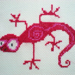Könnte beinhalten: Eine rosa Kreuzstich-Stickerei eines Leguan mit einem eingerollten Schwanz und großen Augen.