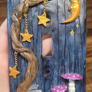 May include: A dark blue rectangular light switch plate with a wood grain texture. It features a sculpted brown tree branch with three hanging gold star charms, a gold crescent moon, two purple mushrooms with white spots, and clear crystals.