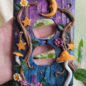 May include: A rectangular light switch cover plate featuring a purple and blue wood-grain texture, adorned with a sculpted crescent moon, orange mushrooms, green leaves, flowers, and hanging gold star charms. The text mysticclaystudios is visible.
