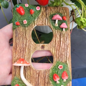 May include: A rectangular light switch cover plate with a textured brown wood grain finish. The plate features raised green moss patches decorated with small red strawberries, pink flowers, and various pink and red mushrooms. The text mysticclaystudios is printed at the bottom.