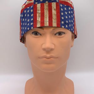 May include: A patterned cap featuring the American flag design. The cap has a blue field with white stars and red and white stripes. The fabric has a slightly distressed, vintage look.