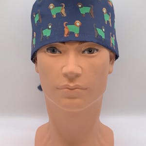 May include: A navy blue surgical scrub cap featuring a repeating pattern of brown dogs wearing green sweaters and face masks. The cap is tied at the back and is designed for medical professionals or pet lovers.