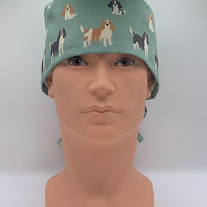 May include: A teal surgical scrub cap featuring a repeating pattern of Cavalier King Charles Spaniel dog illustrations in various colors. The cap is tied at the back and is displayed on a mannequin head.