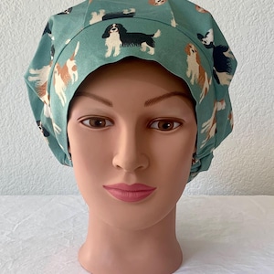 May include: A teal blue surgical scrub hat with a pattern of Cavalier King Charles Spaniels. The hat has a gathered top and a wide elastic band.