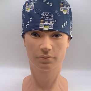 May include: A navy blue scrub cap featuring a repeating pattern of medical vials, syringes, and the text "SWEET dreams ARE MADE OF" interspersed with white "Z" sleep symbols and cloud outlines. The fabric has a dark blue base color throughout.