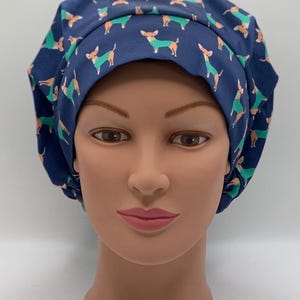 May include: A navy blue scrub cap featuring a repeating pattern of small dogs in orange and green. The cap is designed to cover the head and hair, commonly used in medical or veterinary settings. The fabric appears to be cotton or a similar material.