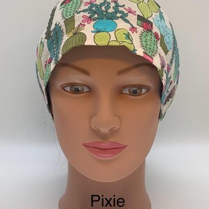 May include: A scrub cap with a cream background featuring a repeating pattern of various green and blue cacti in pots, accented with small pink flowers. The word Pixie is printed in black text at the bottom of the mannequin head display.