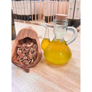 May include: A clear glass cruet filled with yellow liquid, featuring a curved spout and a cork stopper. Beside it, a wooden scoop holds a pile of pecans. The items are arranged on a light-colored wooden surface.